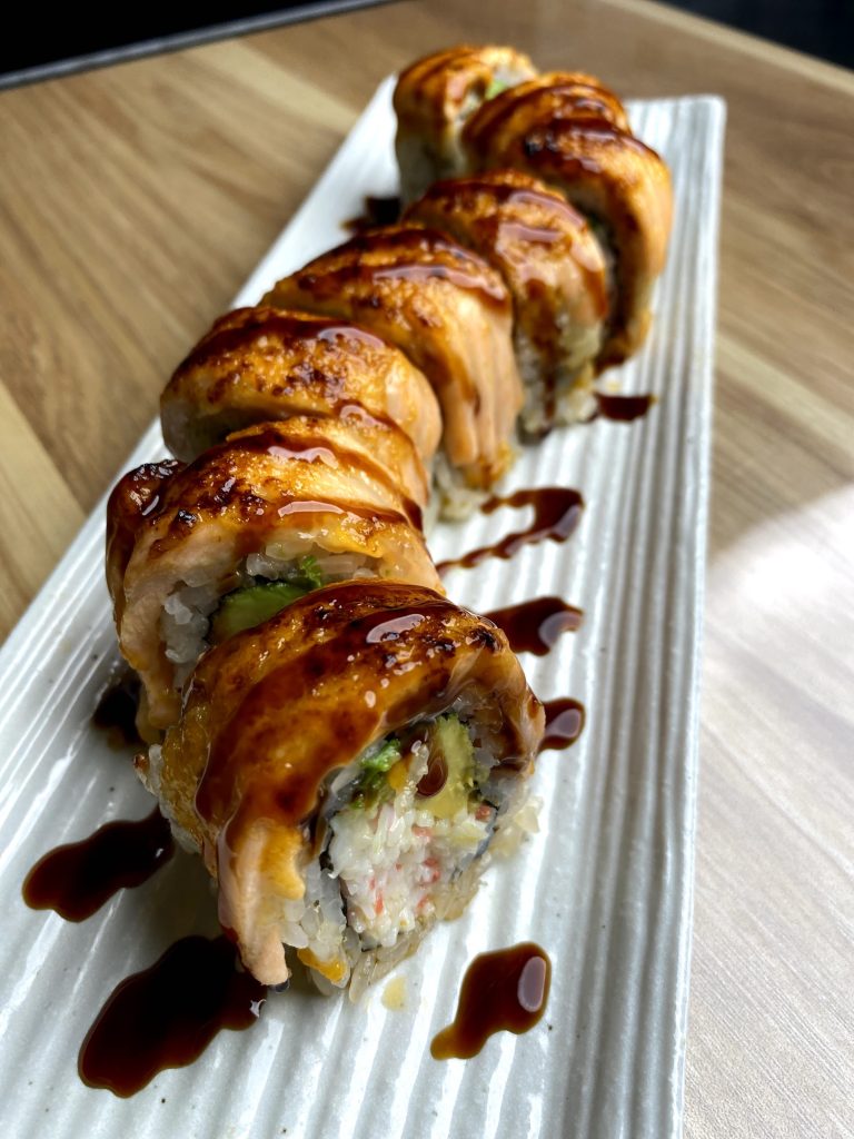 golden dragon roll off white rice rilled with crab and avocado, topped iwth slices of fish and drizzles with brown sauce on white plate on table